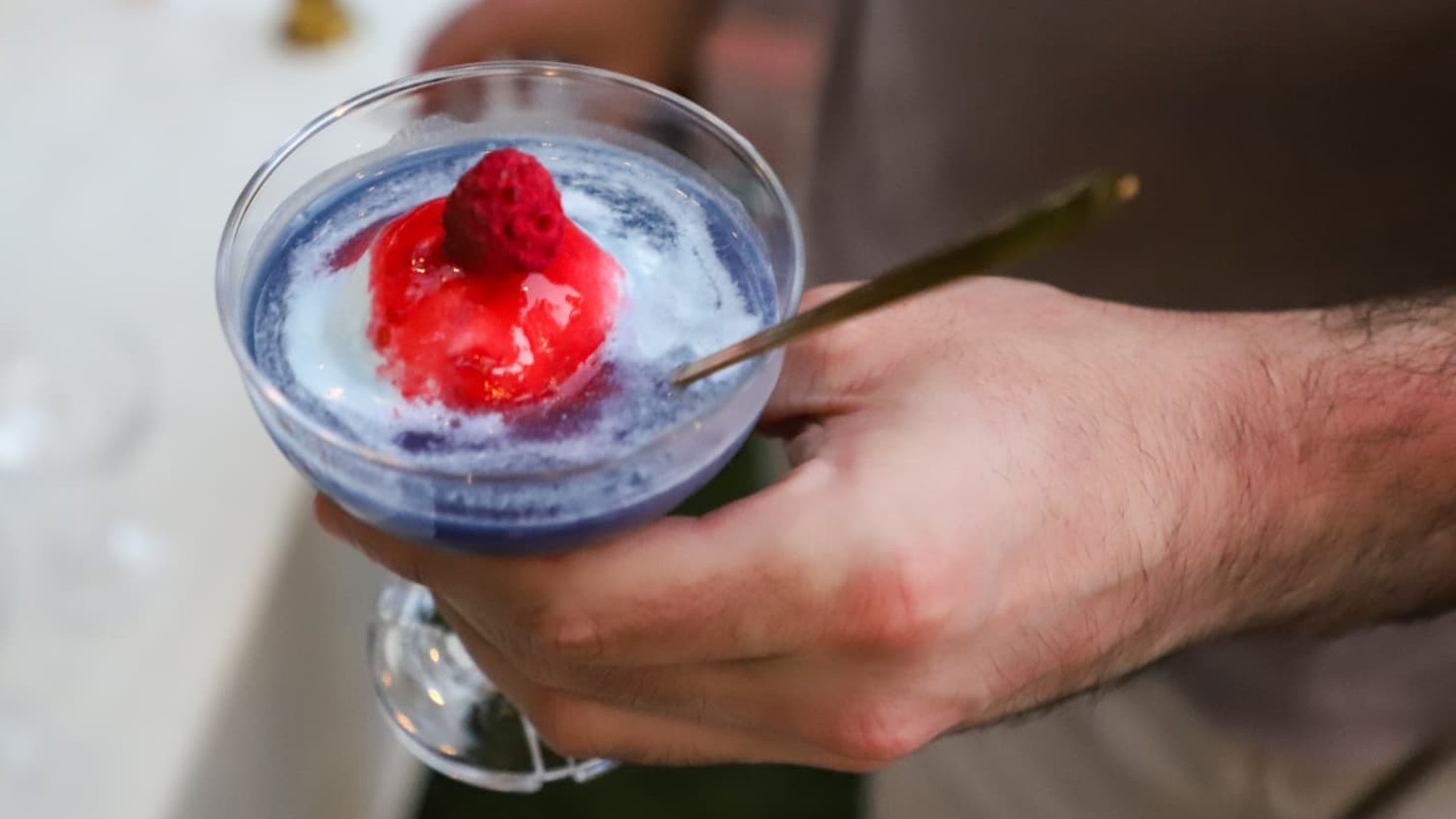 A hand holding a butterfly blue coupe topped with a raspberry and a gold spoon